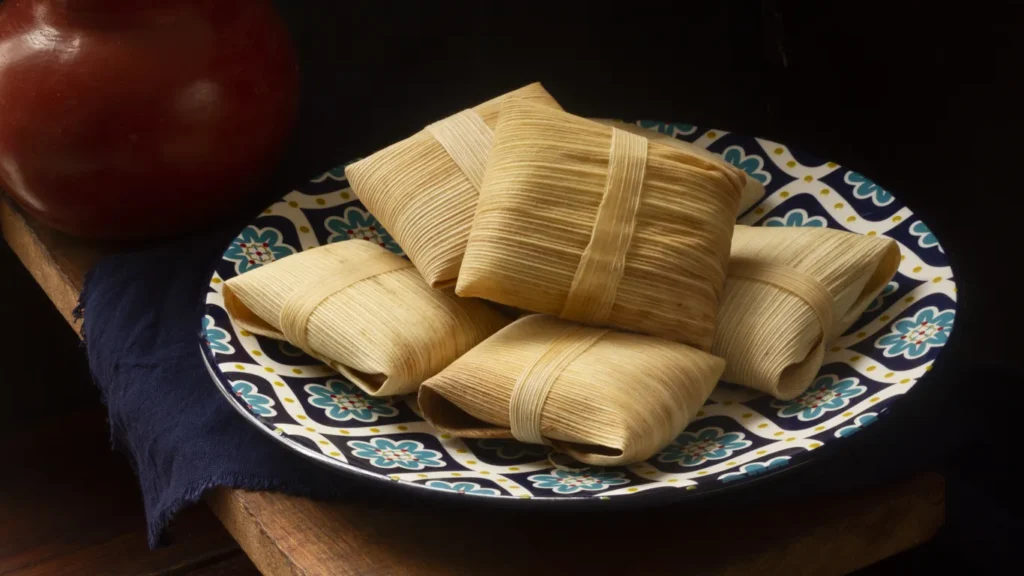 Tamal criollo peruano recién cocido desenvolviendo su hoja de plátano, mostrando la masa dorada cremosa con relleno de cerdo, acompañado de salsa criolla y aceitunas sobre mesa de madera rústica.